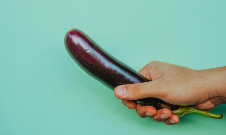 Mão de homem segurando beringela pequena para mostrar como bater punheta sem gozar.