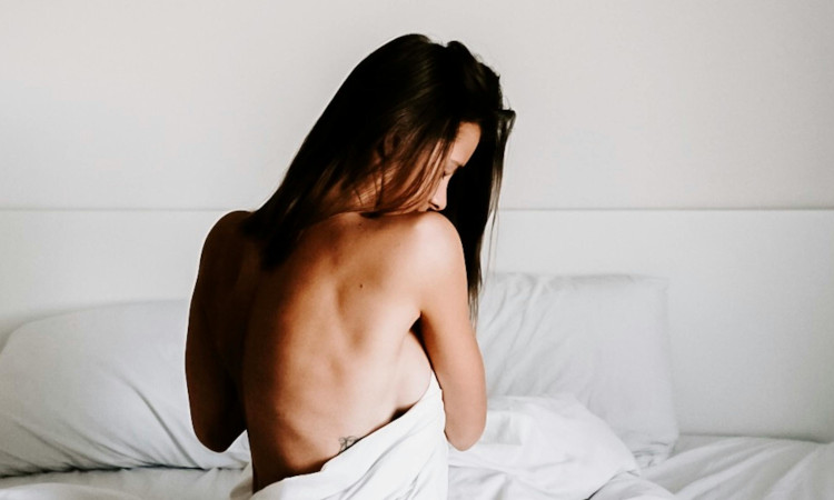 Mulher morena na cama, envolvida num lençol a esconder os seios, sem roupa vestida, e a pensar em como pedir nudes.