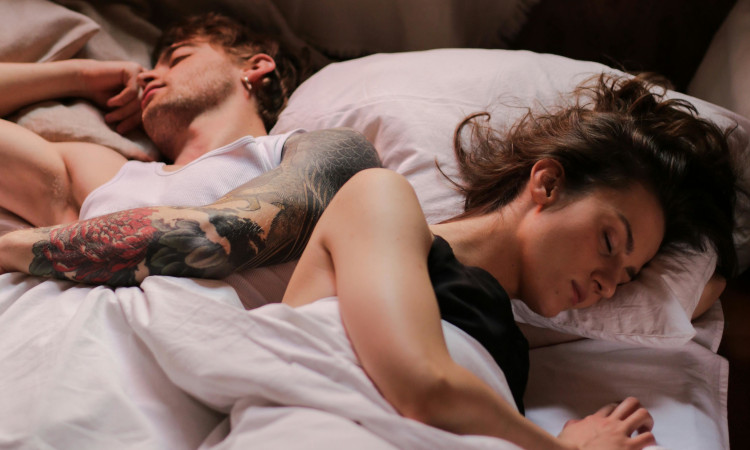 Casal jovem de homem e mulher deitados na cama, cada um virado para o seu lado, e pensando "porque não consigo gozar".