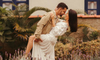 Casal de homem de barba e mulher com vestido branco a sorrirem enquanto pensam em como fazer sexo romântico num campo com flores amarelas e brancas.