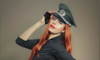 Mulher com cabelo vermelho, lábios pintados de vermelho, chapéu militar na cabeça e camisa preta se preparando para role-play no sexo.